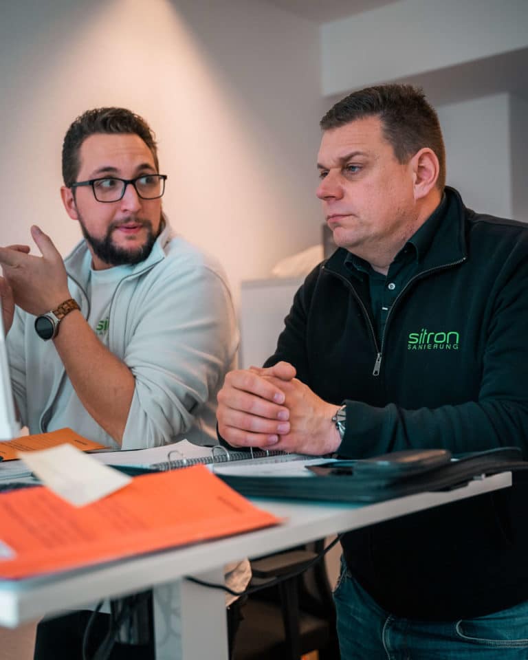 Zwei Mitarbeiter von Sitron mit Arbeitskleidung stehen an einem Schreibtisch und schauen gemeinsam auf Computerbildschirme, um einen Auftrag zu besprechen.